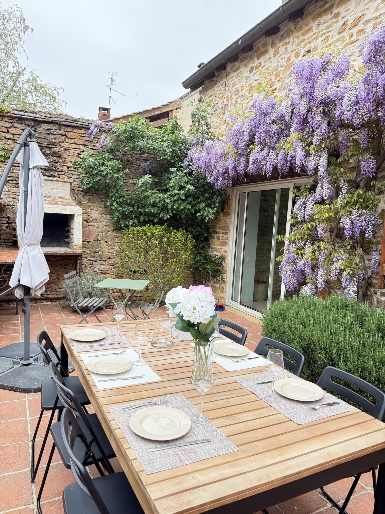 Terrasse avec table dressée devant la façade en pierres couverte de glycine